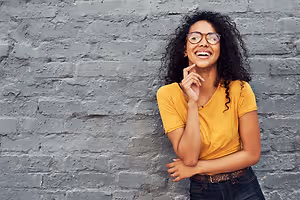 A woman leaning against a brick wall with a yellow shirt and glasses