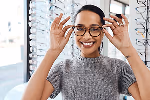 A smiling woman in a gray sweater is adjusting her glasses in front of a glass display of glasses in a store.