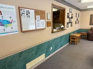 A room with a bulletin board, a desk, and a window with a mirror, as well as a couch and a table with a book on top.
