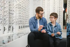 A man and a boy are sitting in an optical shop, and the man is showing a pair of glasses to the boy.
