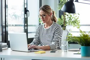 A woman wearing glasses and a striped shirt is sitting on a chair and working on her laptop