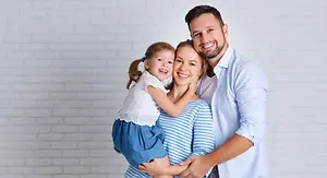 A smiling family with a young girl standing between a man and a woman in a white room
