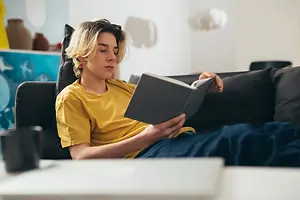 A young man is sitting on a couch and reading a book with a cup and laptop nearby