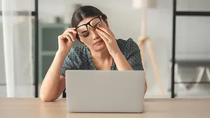 A woman is sitting on a chair in front of a desk, wearing glasses and holding her head, looking at the laptop screen, perhaps in a room, maybe a home office, and behind her is a cabinet and a lamp.