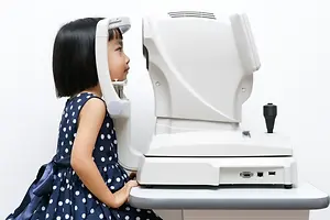 A young girl wearing a polka dot dress sits in front of an eye exam machine in a clinic.