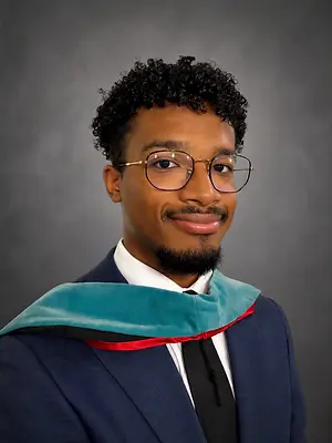 Man wearing a suit and glasses with a graduation gown around his neck smiling for the camera