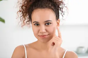 A woman with curly hair wearing a white sleeveless top is touching her cheek with a smile on her face in a room.