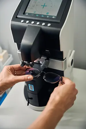 A person's hands are adjusting glasses on an automatic frame measurement machine, a black and white machine with a digital screen displaying measurement details.