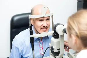 Bald man wearing a blue shirt looks into an eye exam machine while a woman looks on in an office