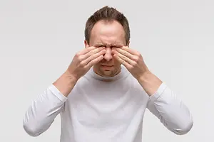 A man in a white shirt with his eyes closed and his hands on his face, looking down.