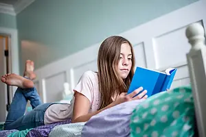 A girl is lying on a bed and reading a book in her bedroom.