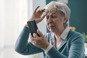 An old woman with white hair is adjusting her glasses while holding a black cellphone