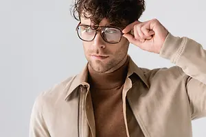A man in a brown turtleneck and a beige jacket adjusts his glasses in front of a white background.