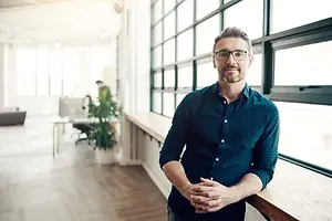 A man with glasses standing in an office room with a window in the background.