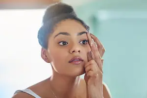 A woman is in a room with a window, touching her eye and looking at her reflection in a mirror.
