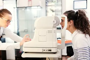 Two women are in an eye clinic, with one woman using a white machine for eye examination.