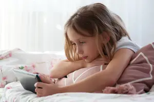 A girl lying on a bed with a pillow and using a tablet in a bedroom