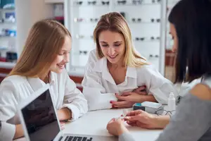 Three women in white lab coats are smiling and sitting around a desk in a room with shelves of glasses. The woman on the left is holding a laptop, and the woman in the middle is holding a small bottle. The woman on the right is holding a piece of paper, and the woman in the middle is touching her stomach.