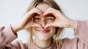 A woman is forming a heart shape with her hands over her eyes and smiling at the camera