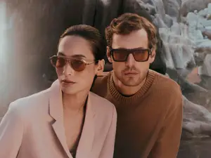 A man and woman stand together wearing sunglasses and light sweaters, with a rocky cliff in the background.