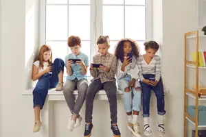 Five children are sitting on a window sill while looking at their phones.