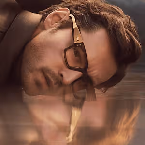Man lying down in a river with his head turned to the side wearing glasses and a coat