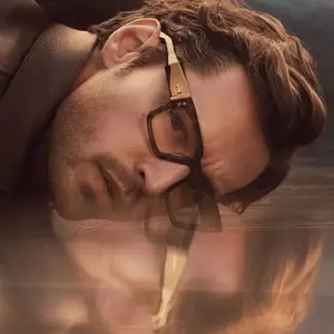Man lying down in a river with his head turned to the side wearing glasses and a coat