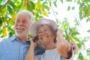 An elderly couple with a woman smiling and placing her hand on her head while the man places his hand on her shoulder in an outdoor area under a tree with leaves and a bright sky