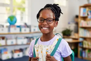 A young girl with glasses and a backpack smiles in a classroom with bookshelves in the background.