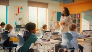 A group of children in a classroom using laptops and being taught by a woman.