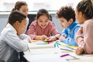 Five children sitting around a desk with pens and pencils, smiling and looking at each other in a classroom