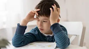A boy looks confused while sitting at a table with a math problem on a piece of paper.