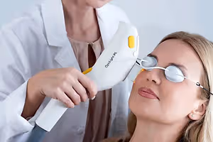 A woman undergoing an eye treatment in a clinic with a professional wearing protective eyewear and using a device labeled 'OptiLight IPL',