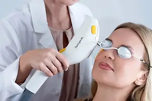 A woman undergoing an eye treatment in a clinic with a professional wearing protective eyewear and using a device labeled 'OptiLight IPL',