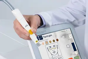 A hand is holding a white and yellow device with the word OptiLight on it, connected to a tablet with a screen showing a diagram of a human face.