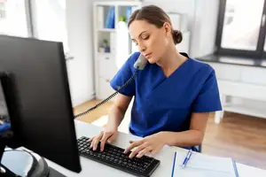 A woman sitting in a chair and using a computer and telephone in an office.