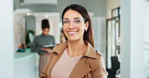 A smiling woman in glasses and a brown coat is standing in a room with a woman reading a book in the background.