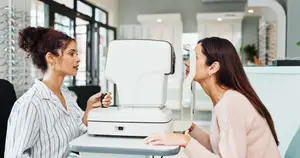 Two women in a medical clinic, one is sitting on a chair while the other is looking into an ophthalmoscope.