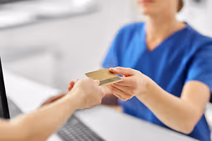 A nurse is holding a credit card in her hand while sitting on a chair in front of a desk with a monitor, keyboard, and mouse.