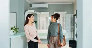 Two women are standing and talking in front of a counter in a room. One of them is holding a handbag and is wearing eyeglasses. The room has a white wall with a glass window, a white counter, and a black chair. There is a potted plant on the counter and an air conditioner on the wall. There are also some frames on display on the wall.