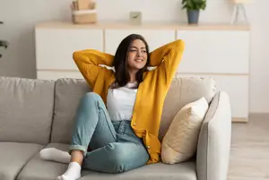 A woman sitting on a couch, resting her arms on her head and smiling while looking at the camera