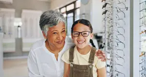 A smiling woman and a girl with glasses are standing in front of a rack of glasses in a store.