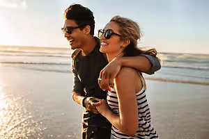 A man and woman smiling on the beach during sunset with the ocean in the background.