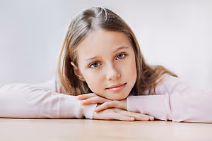 Image of a young girl with blonde hair and freckles leaning on a table with a serious expression.