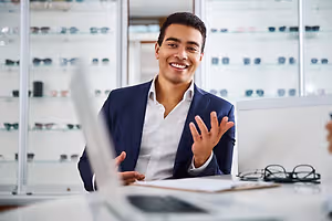 A man is sitting in front of a desk, smiling, and gesturing with his hands.