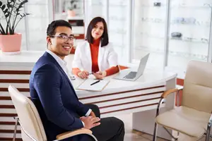 A smiling man wearing glasses is sitting in a chair in front of a desk where a woman is sitting and working on a laptop, a potted plant is on the counter, and glasses are displayed in a glass cabinet.