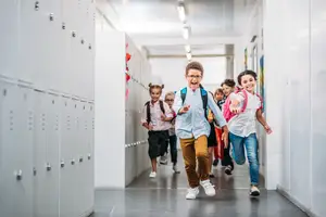 Children run through a school hallway, smiling and excited, with lockers on either side.