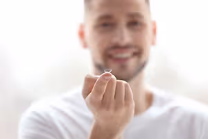 A smiling man in a white shirt is holding a contact lens in his fingers, possibly for a close-up shot.