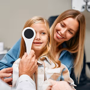 A young girl is having her eyes checked by a doctor while a woman holds her and smiles