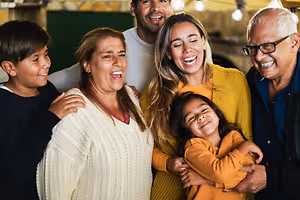 A family of five, including an elderly man, a woman, a girl, a boy, and a man, are smiling and posing for a photo in a room with a brick wall and a green door, along with some hanging lamps and shelves.